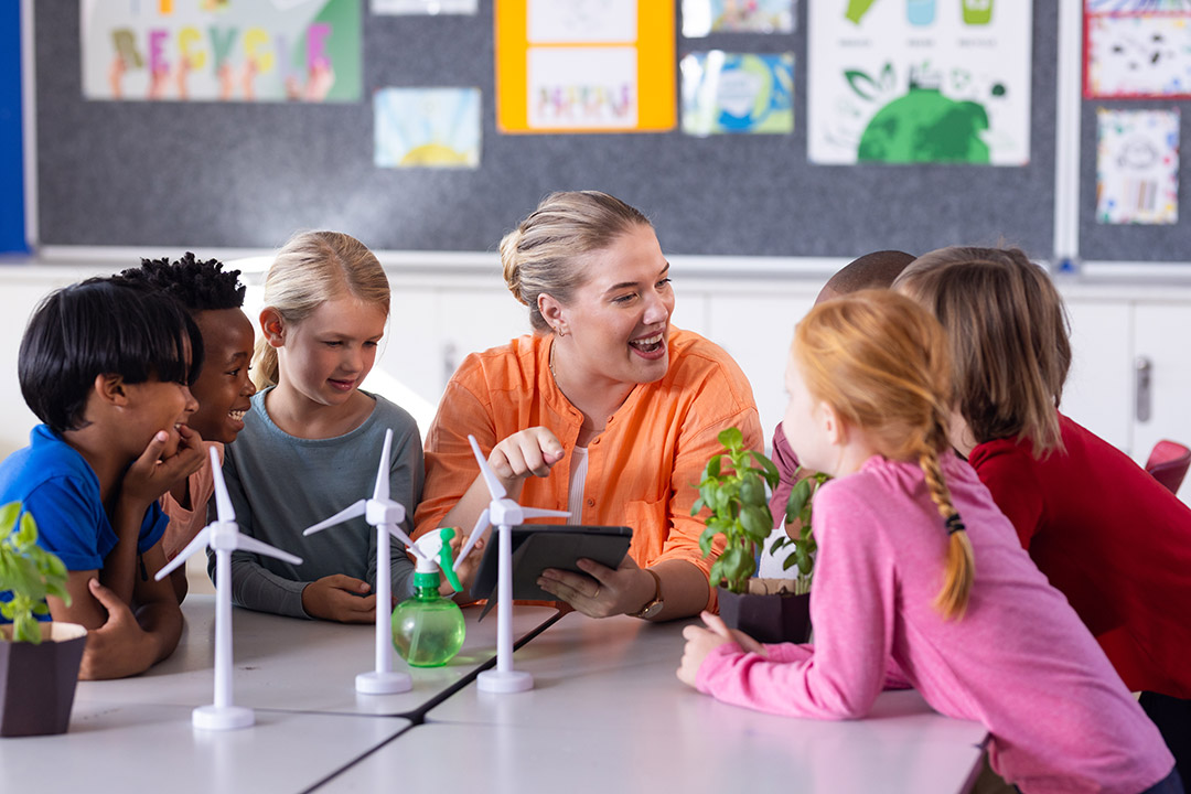 miling teacher in an orange shirt engages with a group of diverse young students around a table with small wind turbines, plants, and a spray bottle in a bright classroom