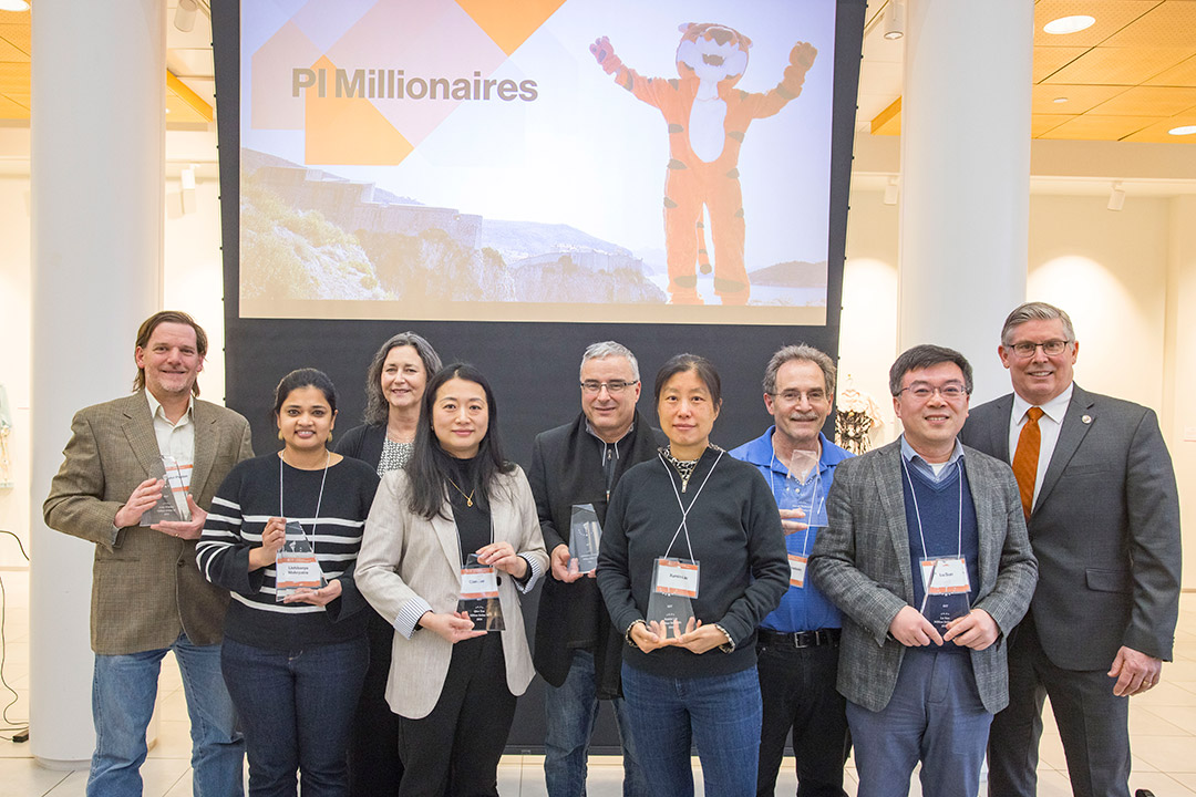 "A group of nine people pose with awards at the PI Millionaires event, standing in front of a screen displaying the event’s name and a tiger mascot. The attendees are dressed in business or smart casual attire, holding glass trophies, and smiling.