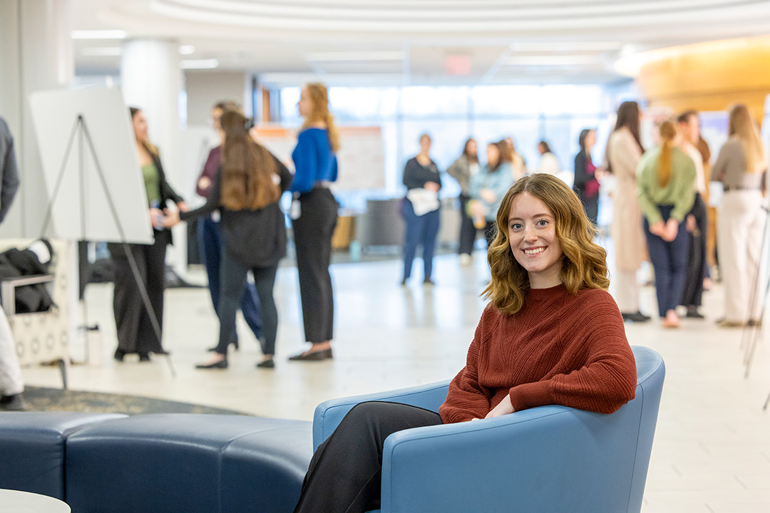 Smiling woman in a red sweater sits in a blue chair in the foreground of a busy networking event with people conversing in the background.