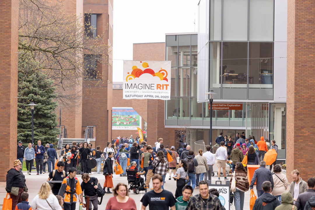 A large group of people walk on campus under an Imagine RIT banner.