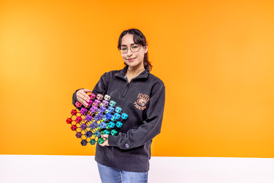 A person holding a colorful geometric hexagon structure against an orange background, wearing an R I T Tigers sweater.