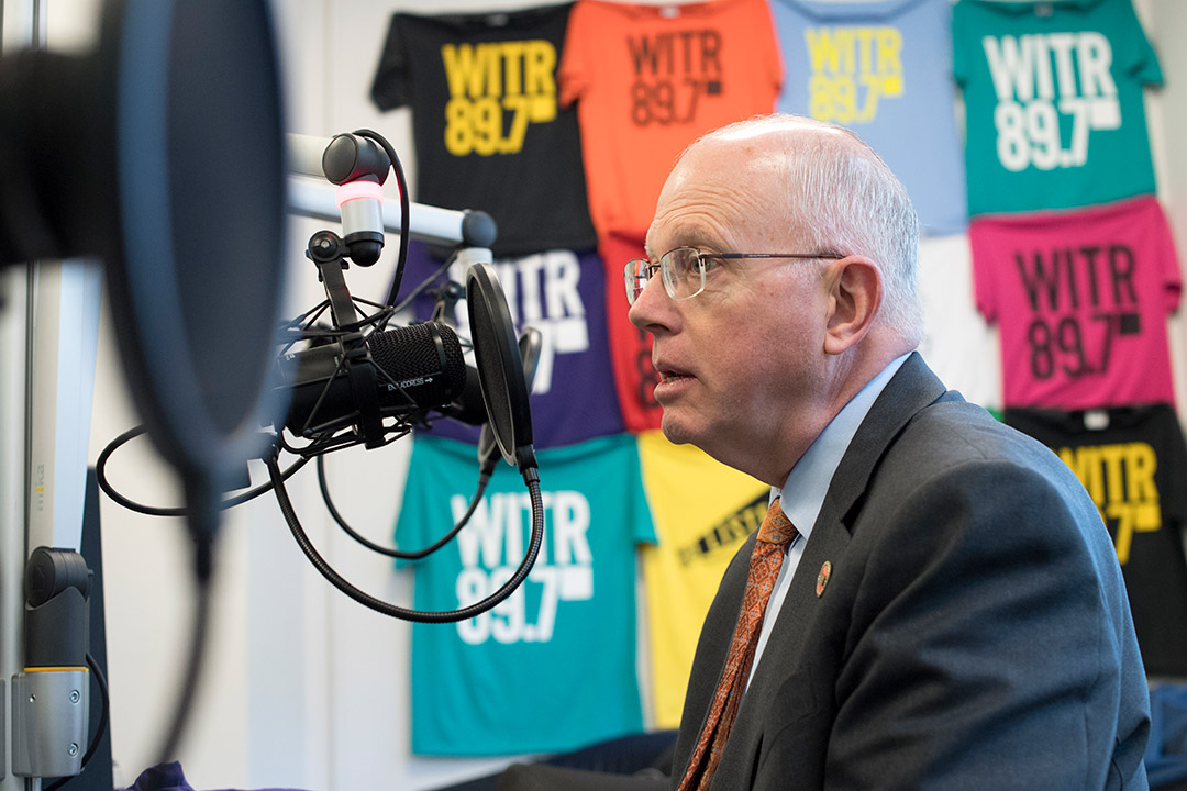 David Munson sits in front of colorful shirts bearing the W I T R logo in a gray suit facing a microphone.