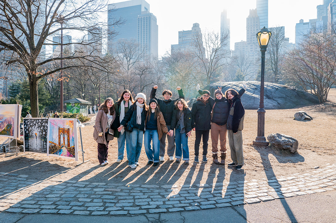 A group of ten people pose in a park under sunny skies, with city buildings in the background.