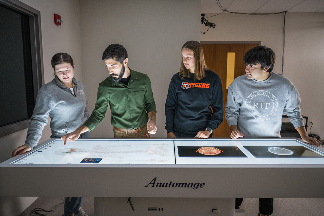 A priofessor and three students stand around a digital cadaver table, which allows for interactive, 3D exploration of the human body.