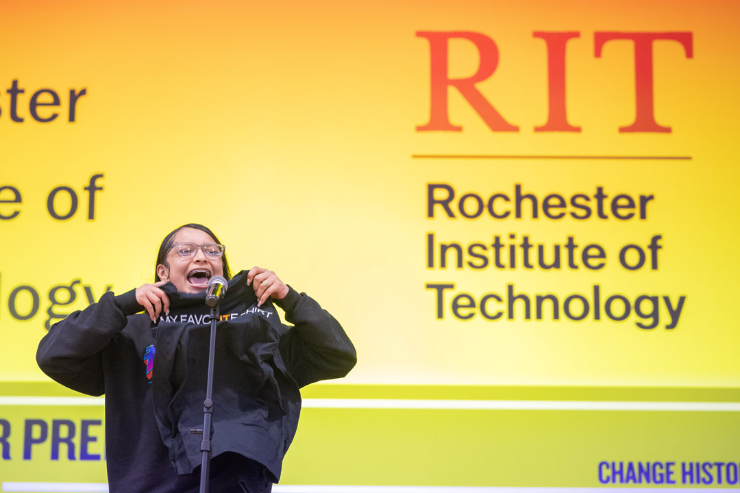 A student opens a black t-shirt in front of a microphone with RIT's logo displayed on a screen in the background.