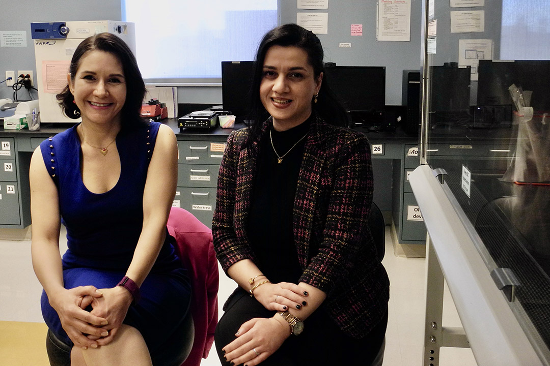 Two women smile while seated in a lab with computers and scientific equipment in the background.