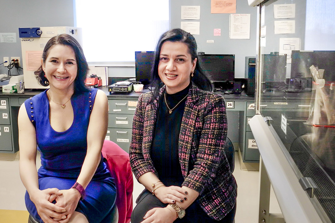 Two women smile while seated in a lab with computers and scientific equipment in the background.