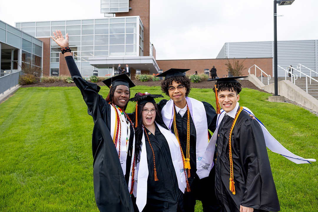 Four graduates in caps and gowns celebrating on a grassy area in front of a modern building.