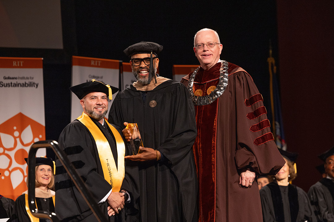 Three men in formal academic attire stand on stage, one holding a glass award.