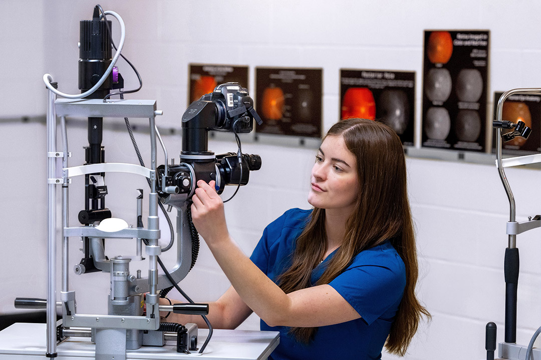 'a female student in a blue shirt works with a camera attached to a microscopy in a white lab room.'