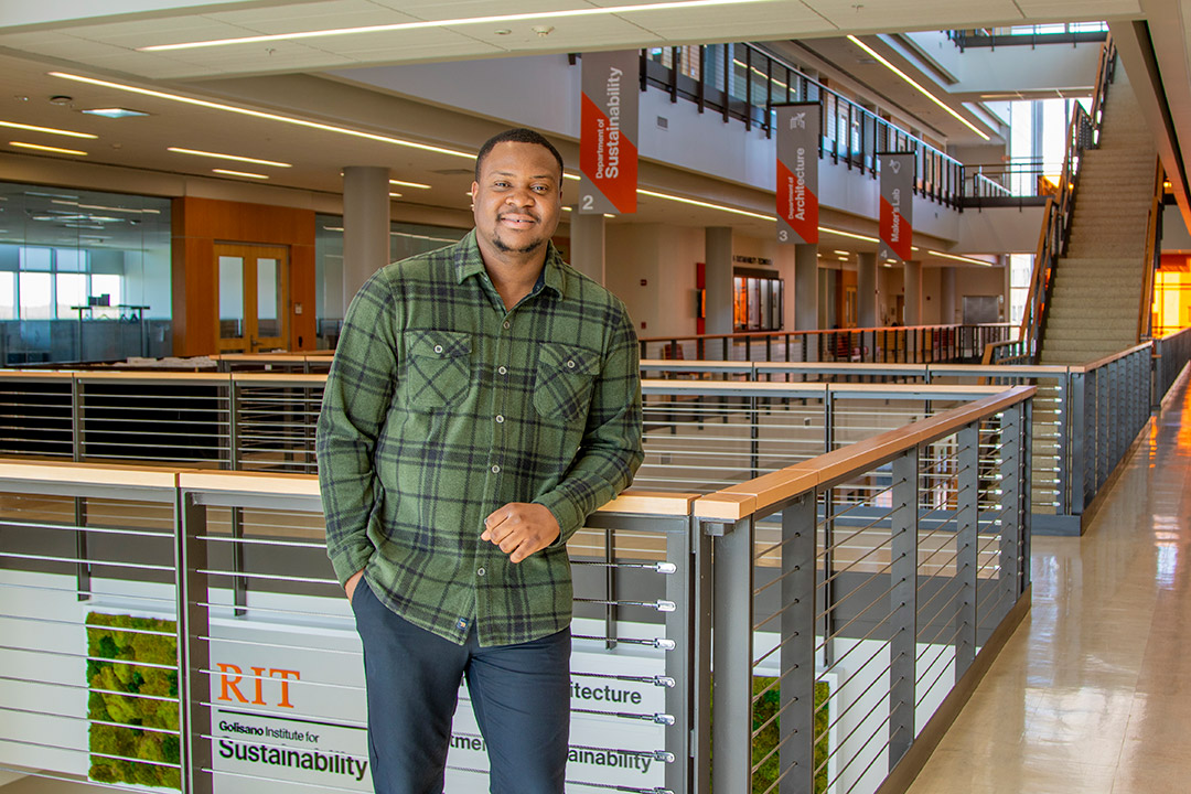 Tunmise Raji stands at a railing in a college building wearing a green shirt and jeans.