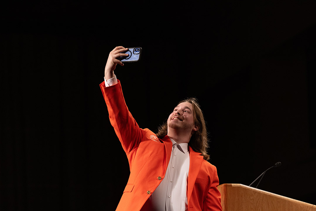 A person in an orange blazer taking a selfie with a phone against a dark background.
