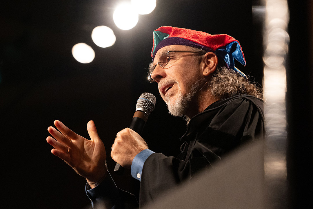 A man wearing a colorful jester hat speaks into a microphone with his hand raised.