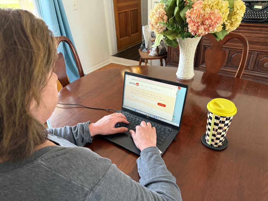 An individual wearing a grey shirt is on a laptop looking at the screen. They have a coffee mug next to them.