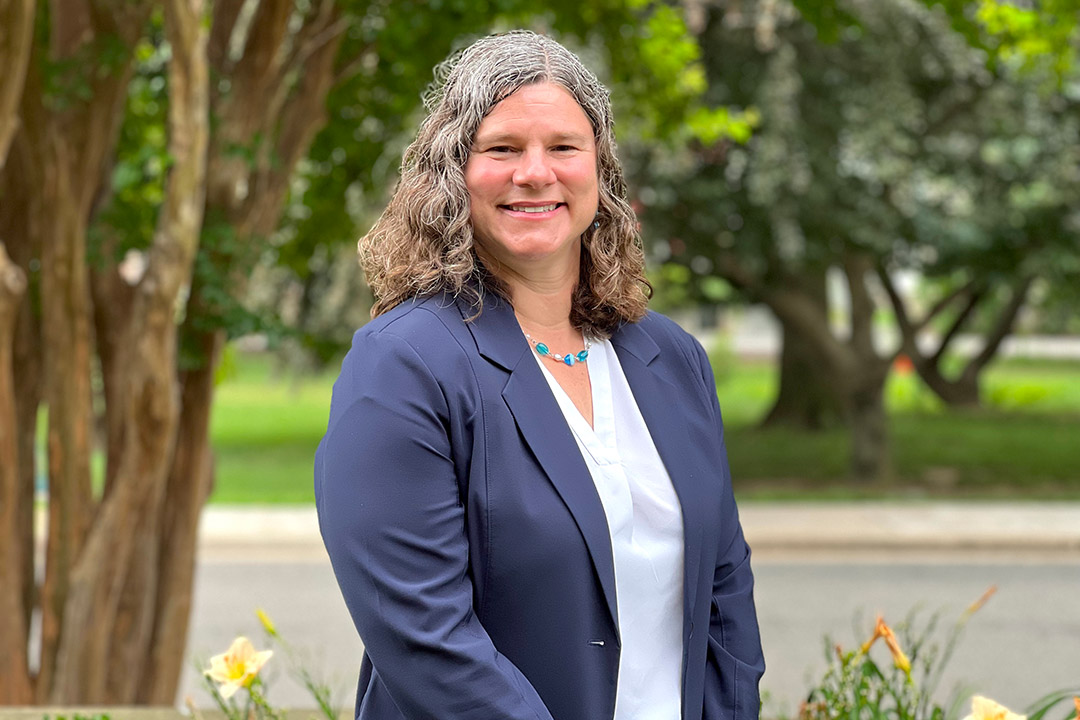 a woman with curly gray hair stands in a park like setting in a navy blue suit.
