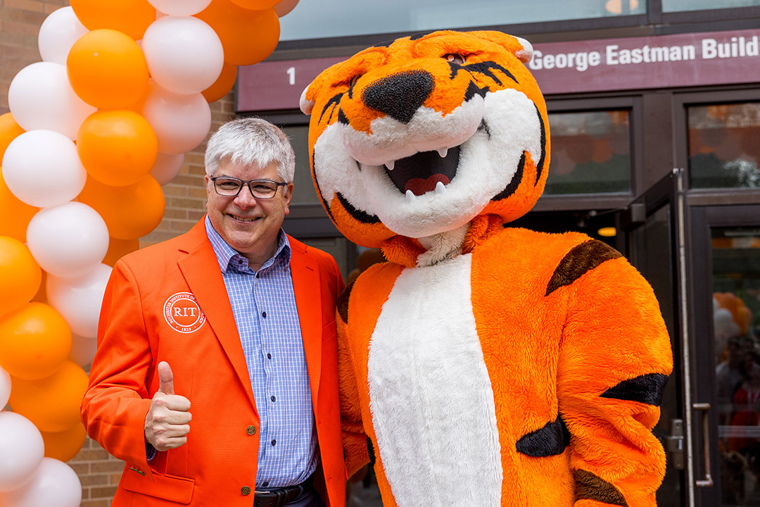 an older man in an orange blazer stands next to R I T's mascot, a tiger.