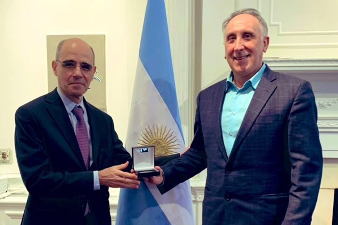 two older men stand next to each other with the man on the left presenting the other with a small box that contains an award. a blue and white flag with a yellow sun is behind them.