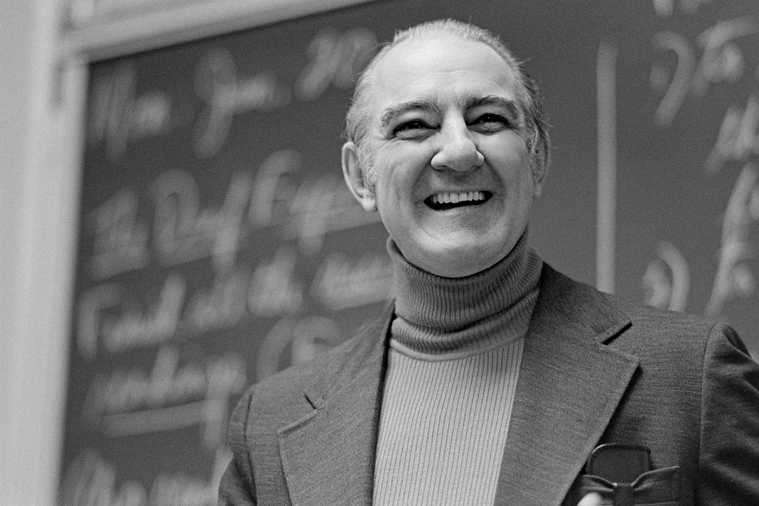 a black and white photograph of an older man dressed in a dark jacket and light turtleneck standing in front of a chalkboard with blurred writing.