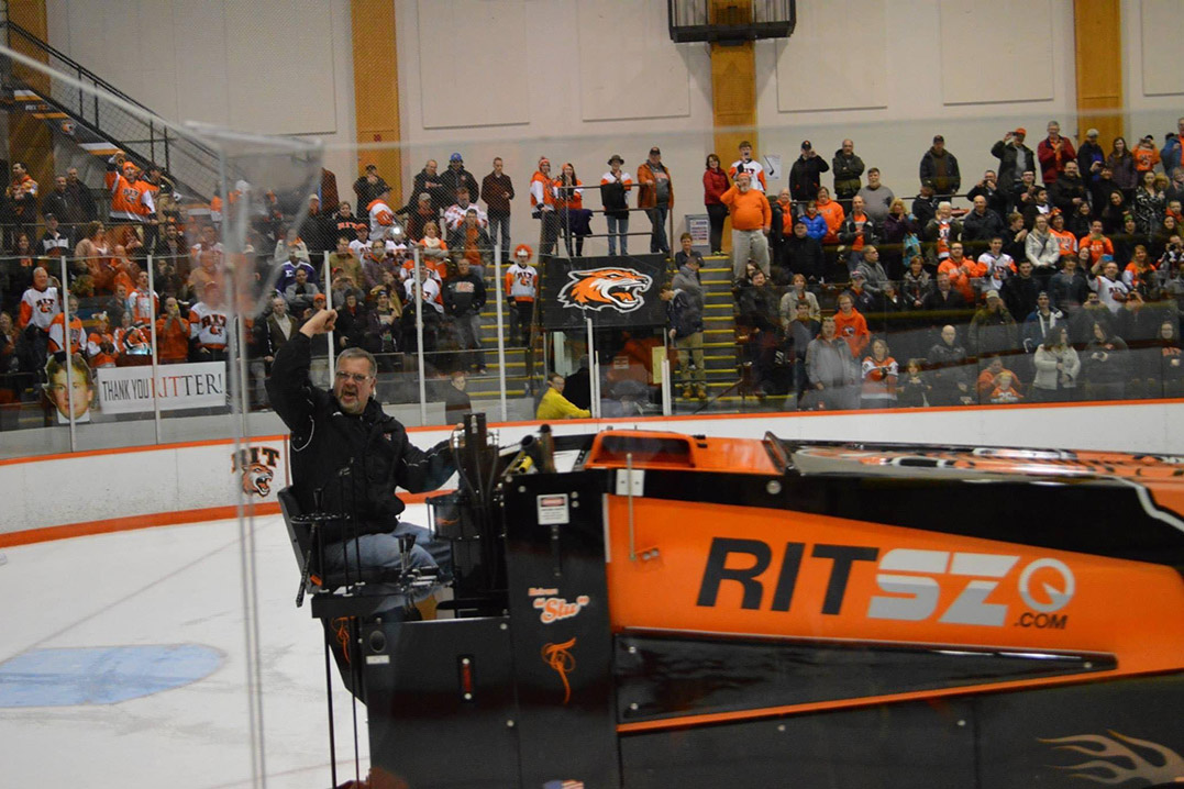 Man proudly driving Zamboni on ice rink during hockey game, waving to cheering crowd.
