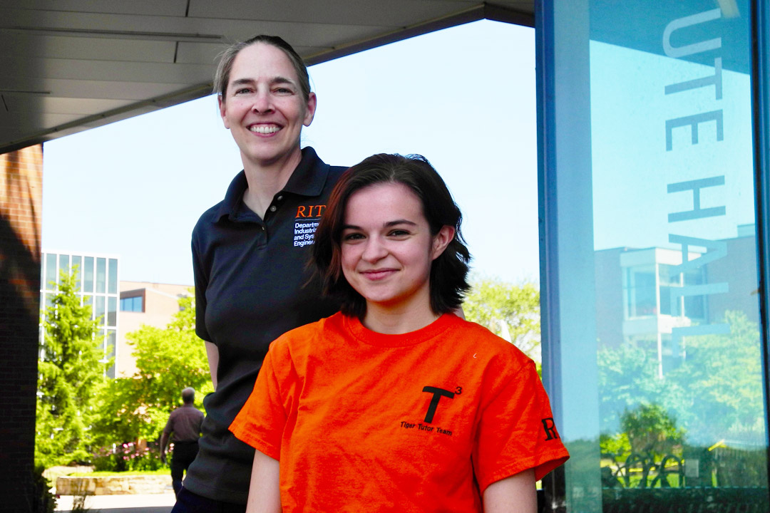 A woman in a black polo shirt stands behind a student in an orange t-shirt.
