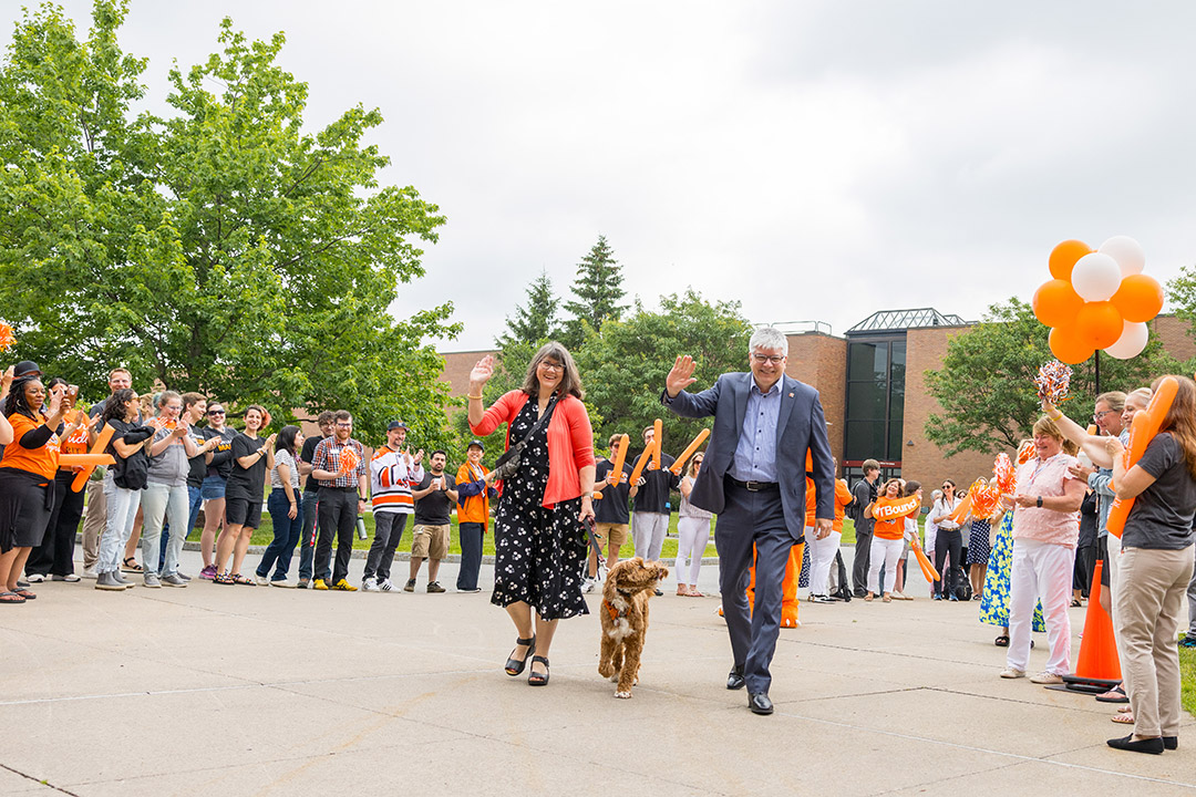 a woman, small dog, and man walk through a crowd of people lining the walkway in an outdoor area.