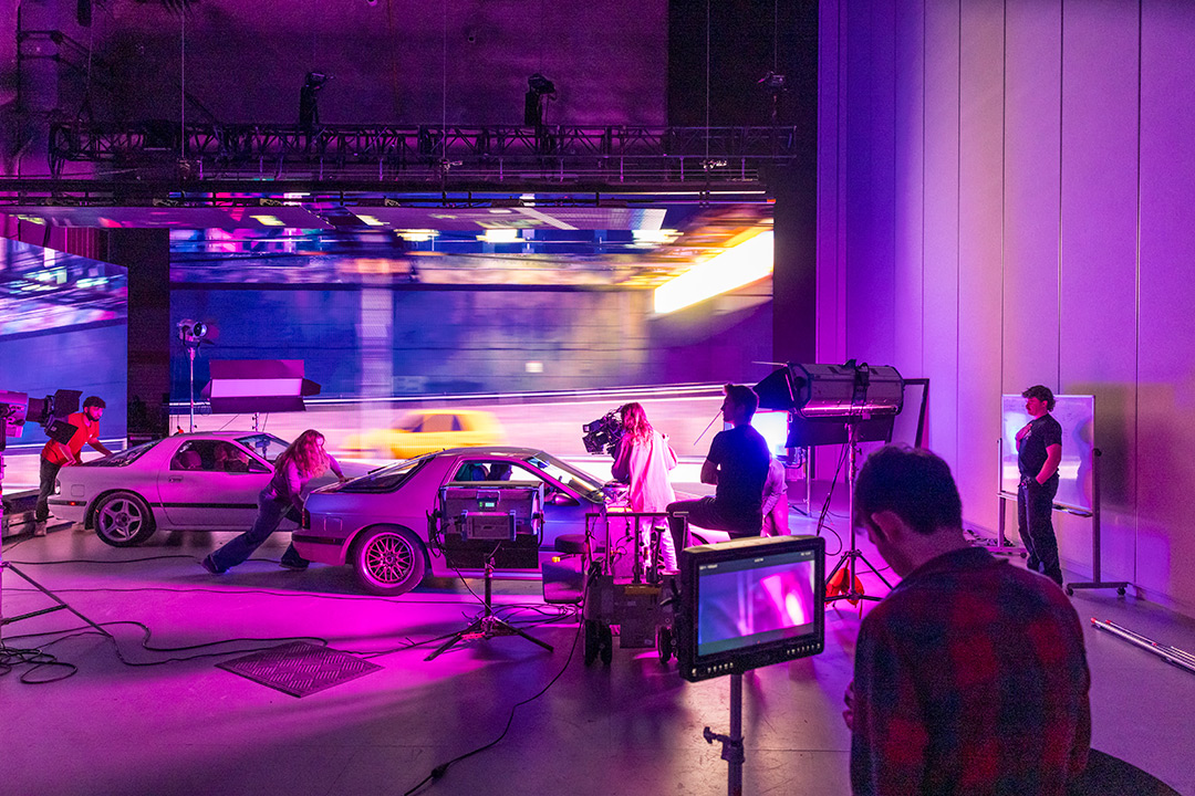 a film crew stages two cars on a purple-lit virtual production set with a moving LED backdrop