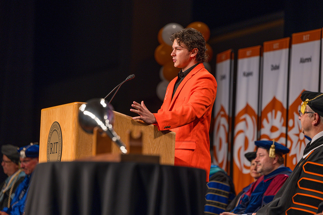 a college age male speaks to a crowd from behind an R I T podium