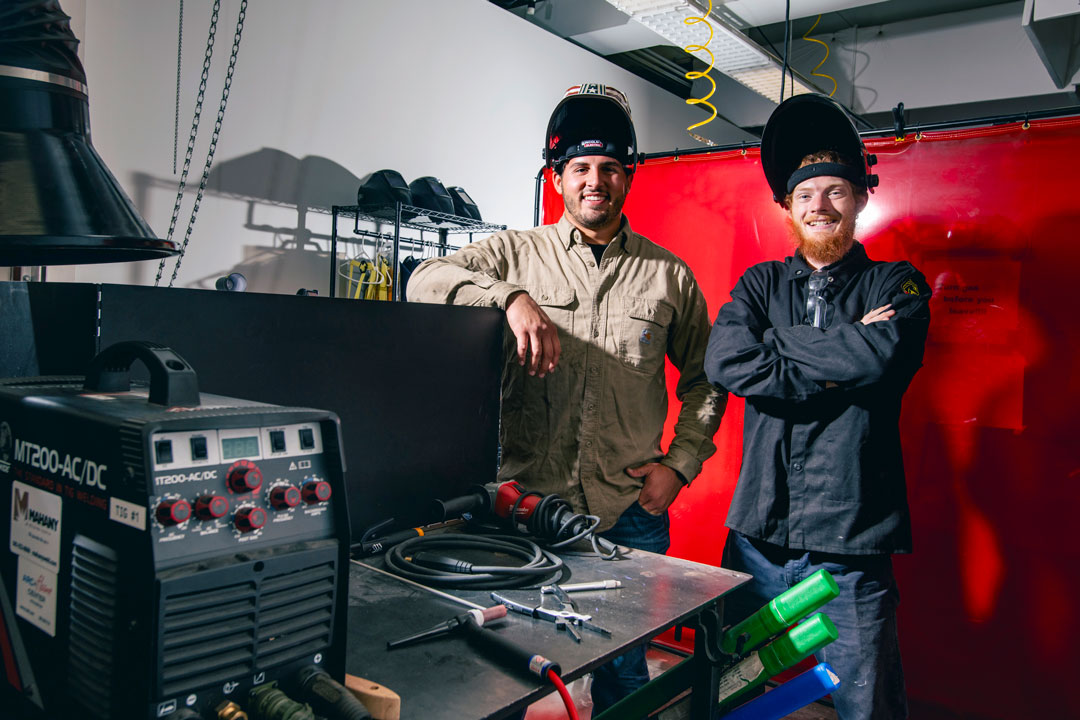 Two people wearing welding helmets stand behind a welding bench with equipment.