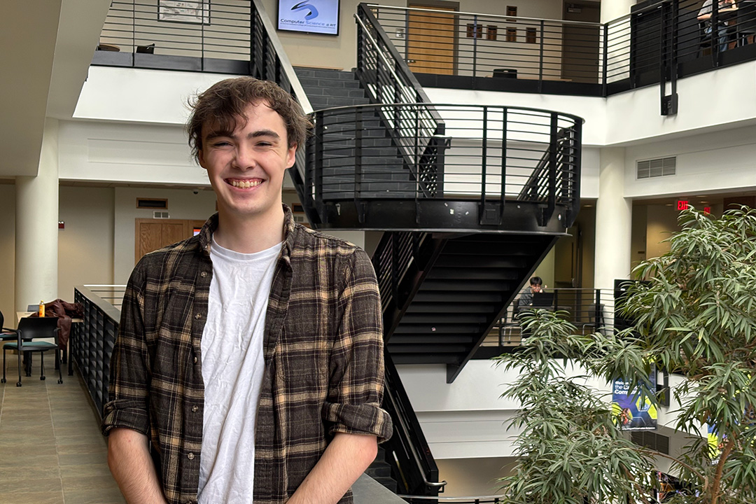student smiling in white shirt and brown plaid shirt stands on 2nd floor of GCCIS atrium
