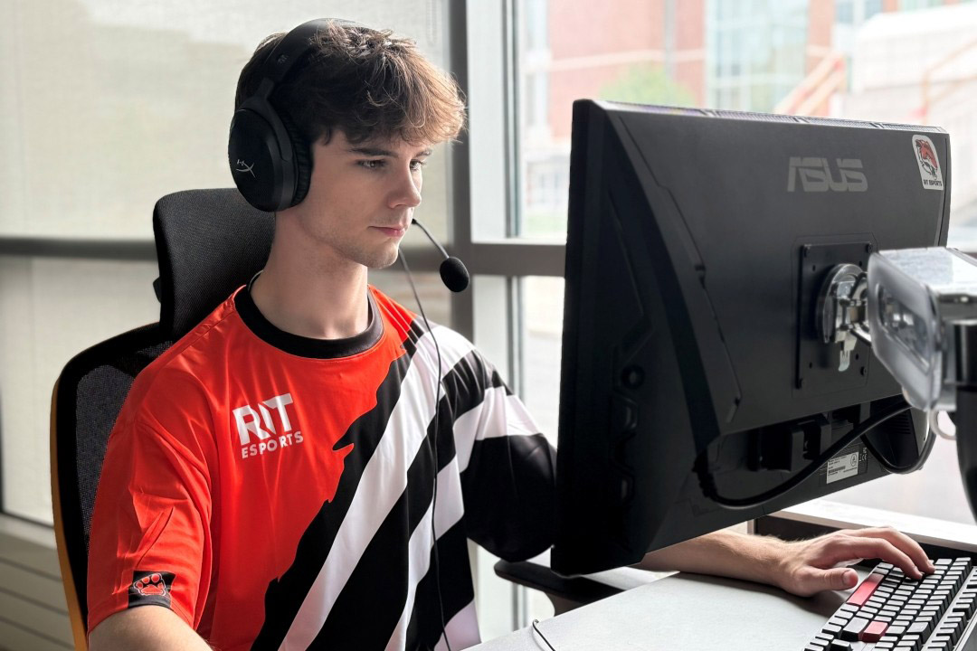 a student in an orange black and white shirt sits at a computer in a well lit area.