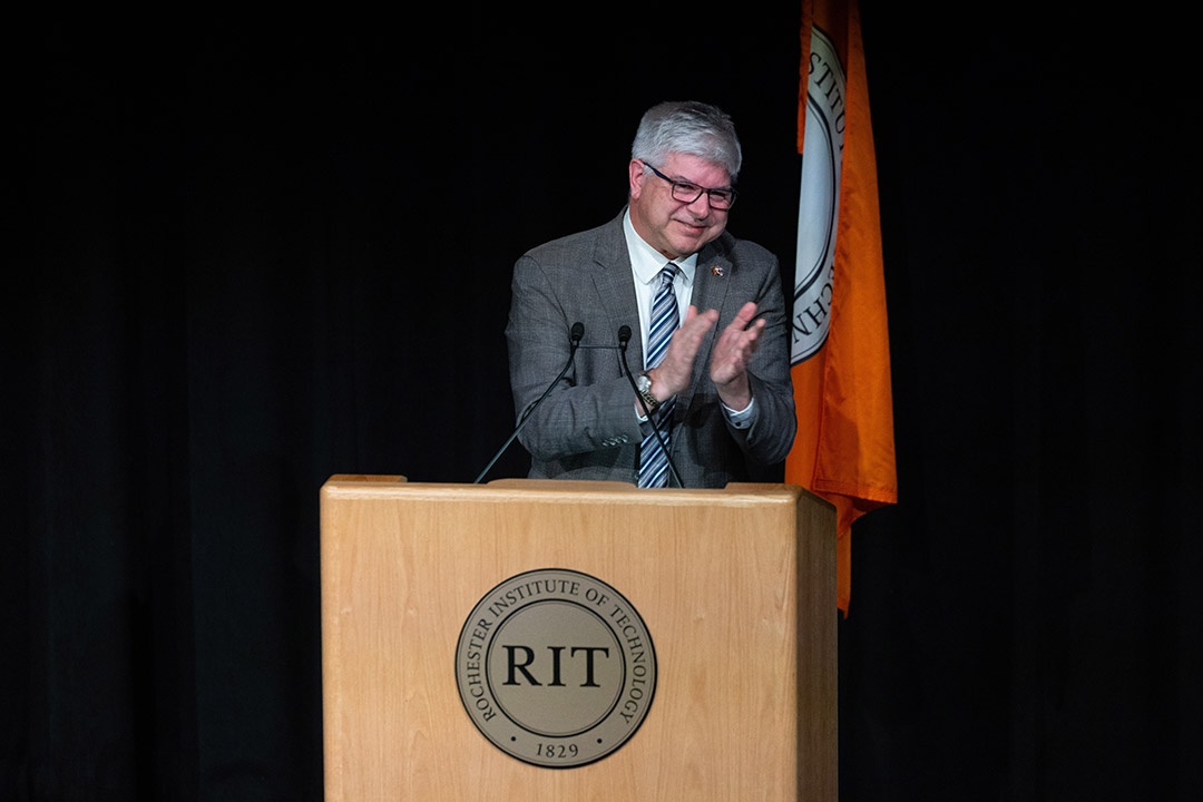 'R I T President Bill Sanders stands on a stage and looks happily at a crowd of people'