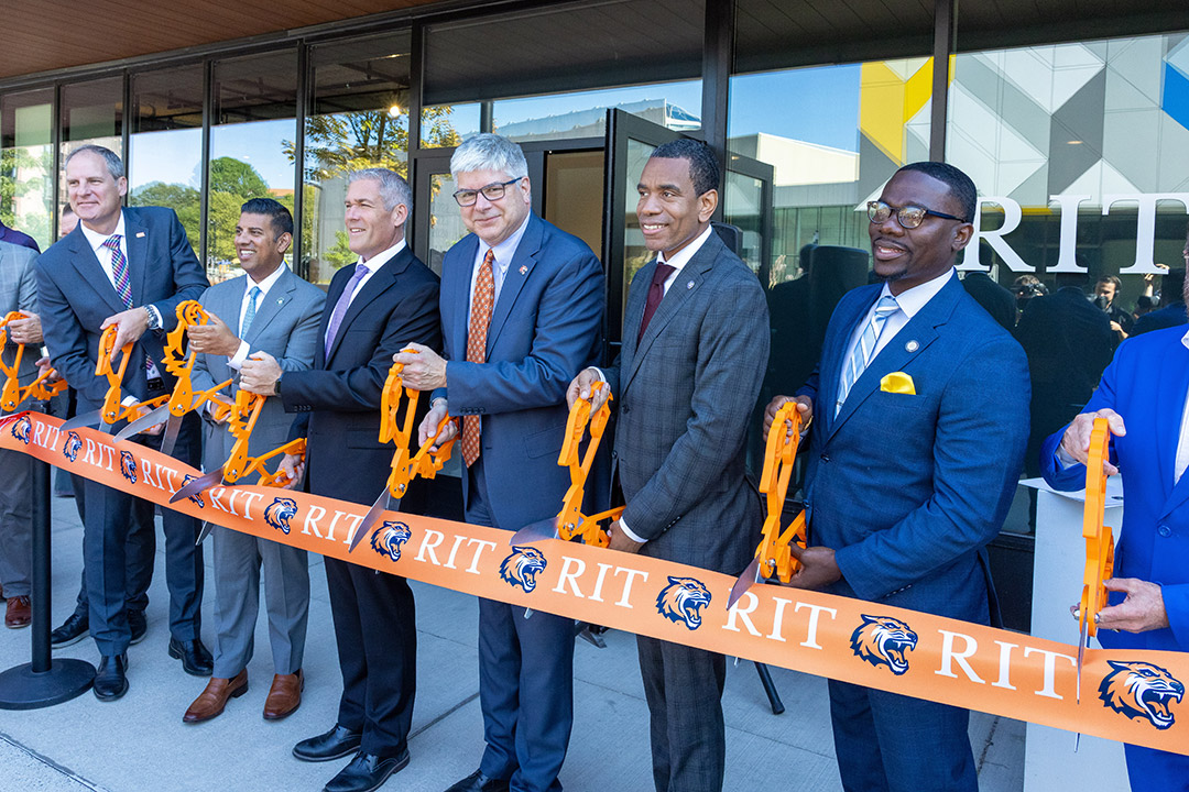 Seven men cut a large R I T printed ribbon in front of an art gallery