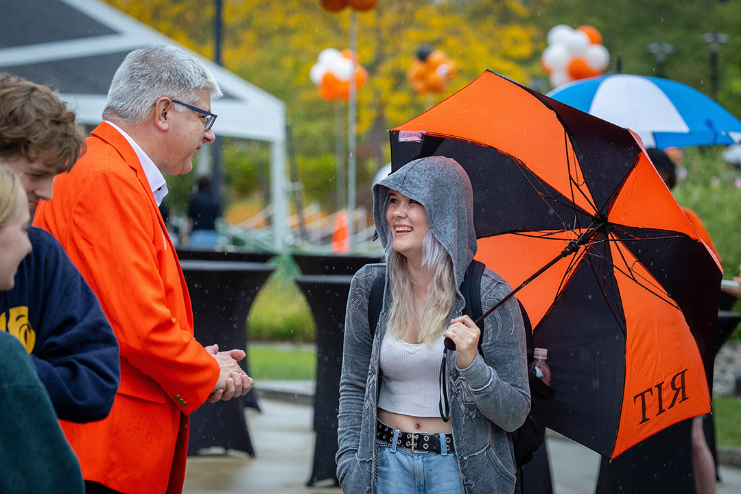 'a man in an orange blazer speaks to a college student carrying an umbrella.'