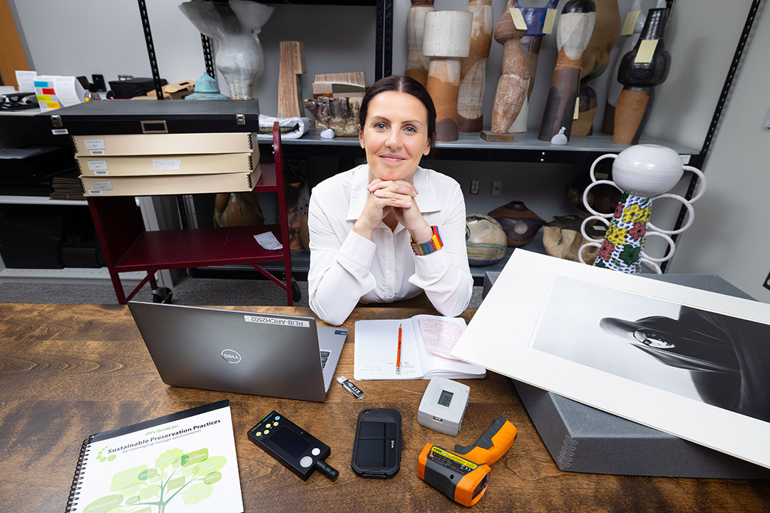 a woman with dark hair sits at a table in an archive environment.