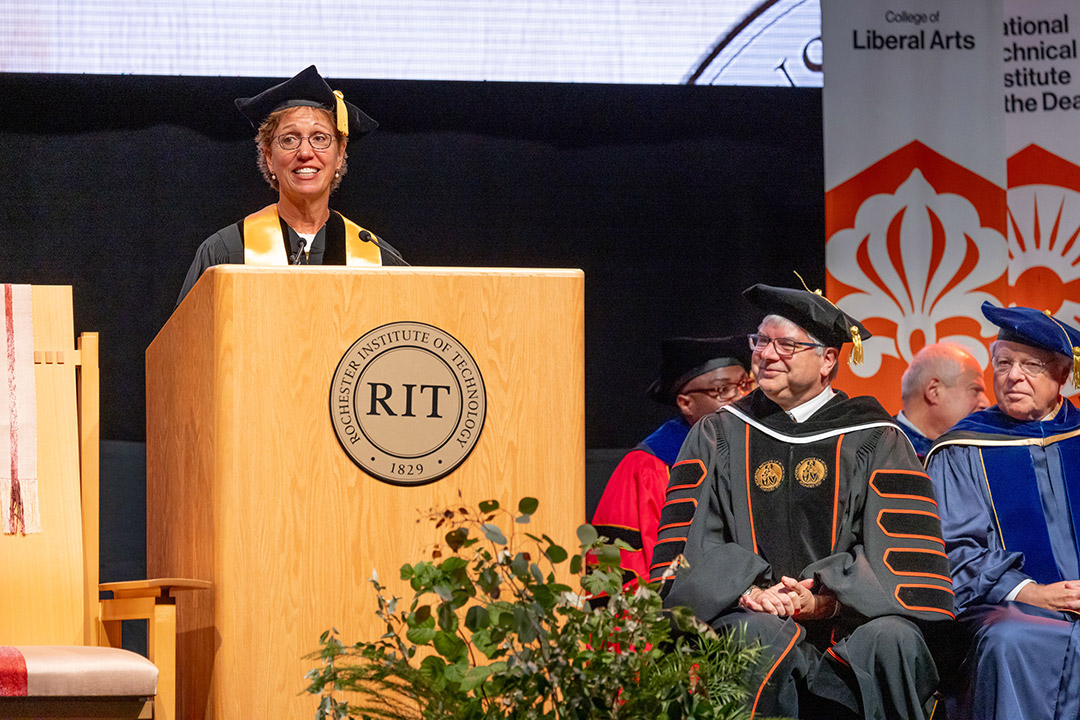 a woman stands behind an R I T podium in univesity regalia.