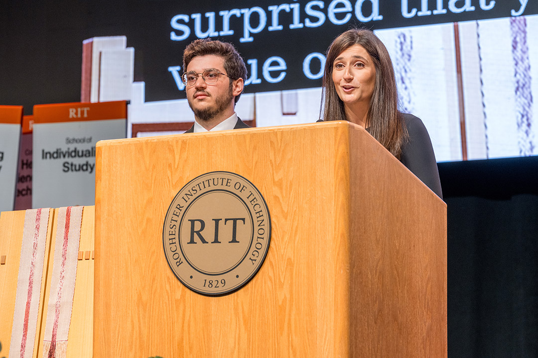 a female stands behind an R I T podium on stage with a presentation displayed on screen next to her. A male stands next to the podium.