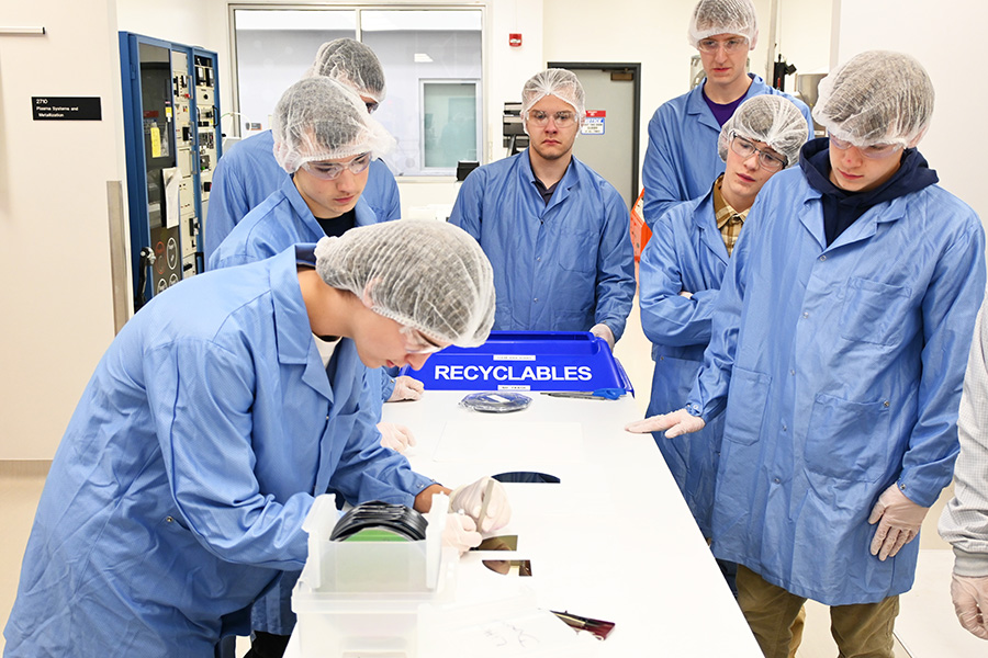 One high school student splits a silicon wafer at a table while six other high school students observe in a clean room facility.