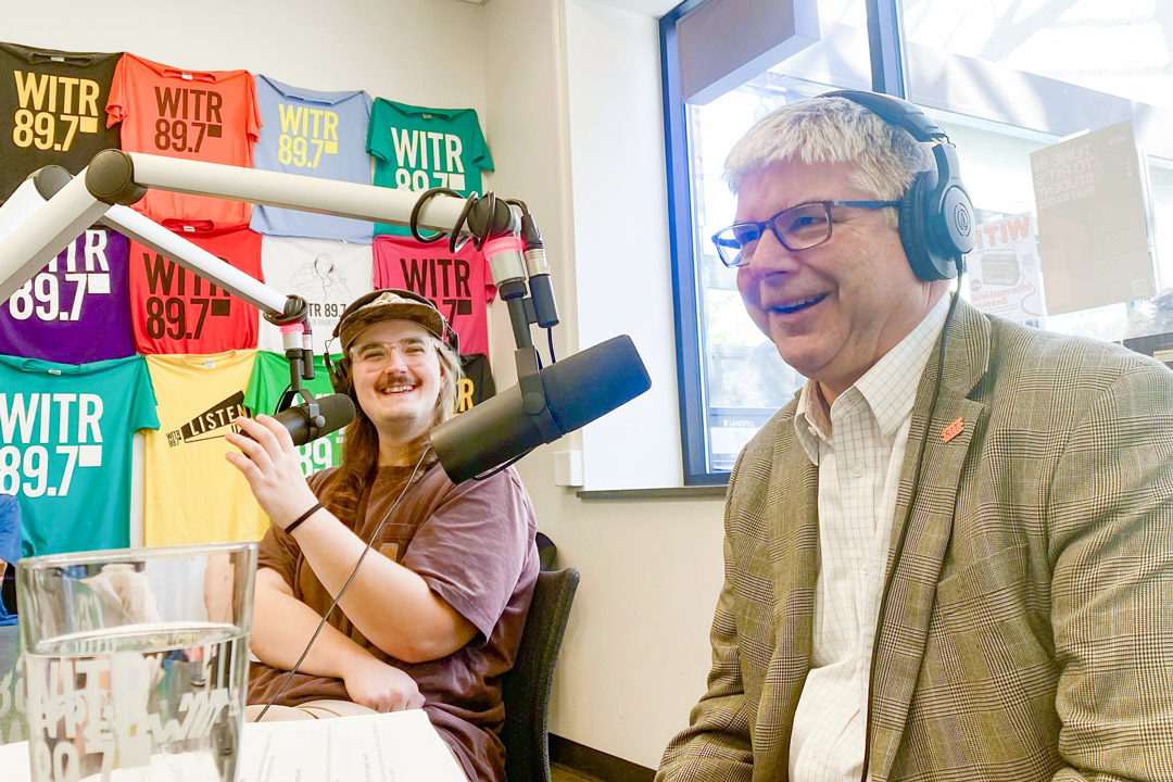 William Sanders sits in a radio studio with a student, both speaking in to microphones.