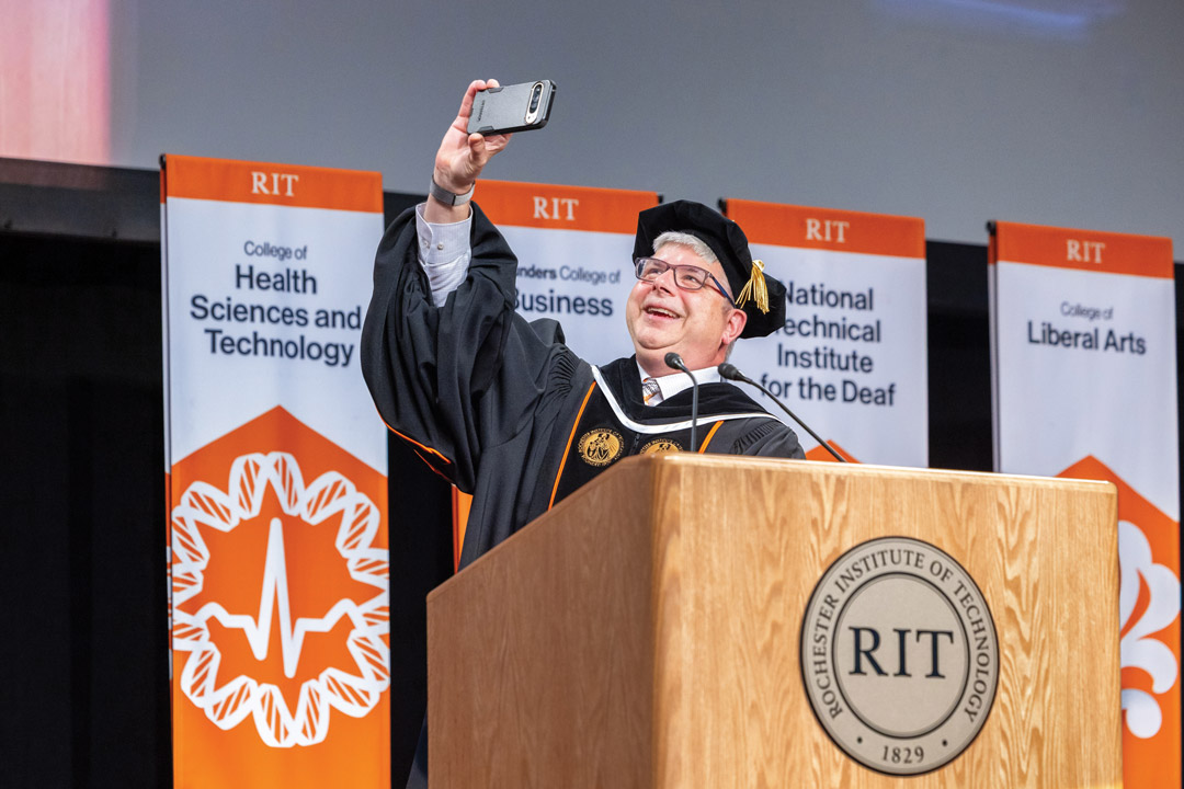 a man in regalia stands at an R I T podium taking a selfie.