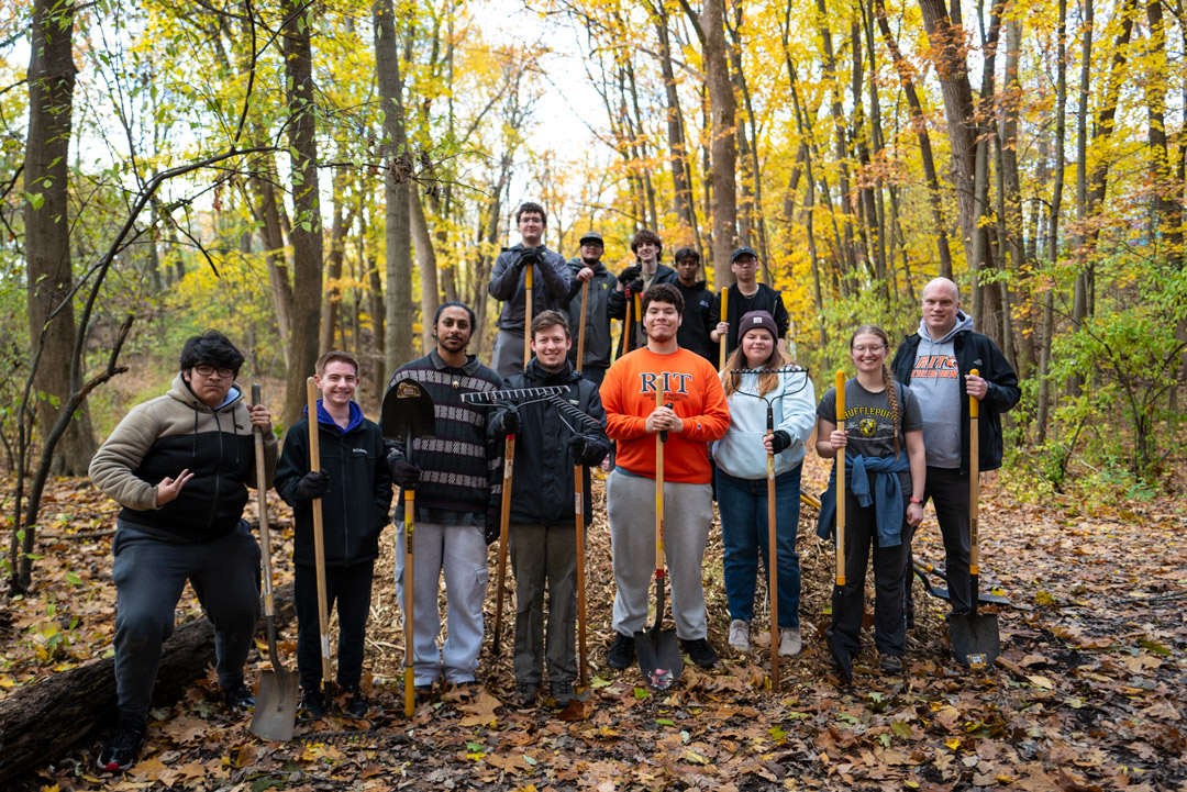 thirteen people stand with yard work tools in a wooded area in the fall.