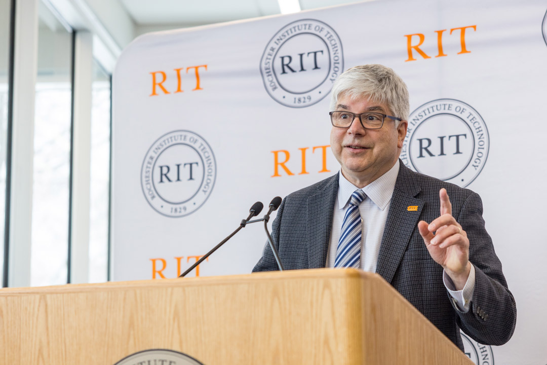 a man in a gray suit stands behind a podium with an R I T logo displayed on a step and repeat behind him.