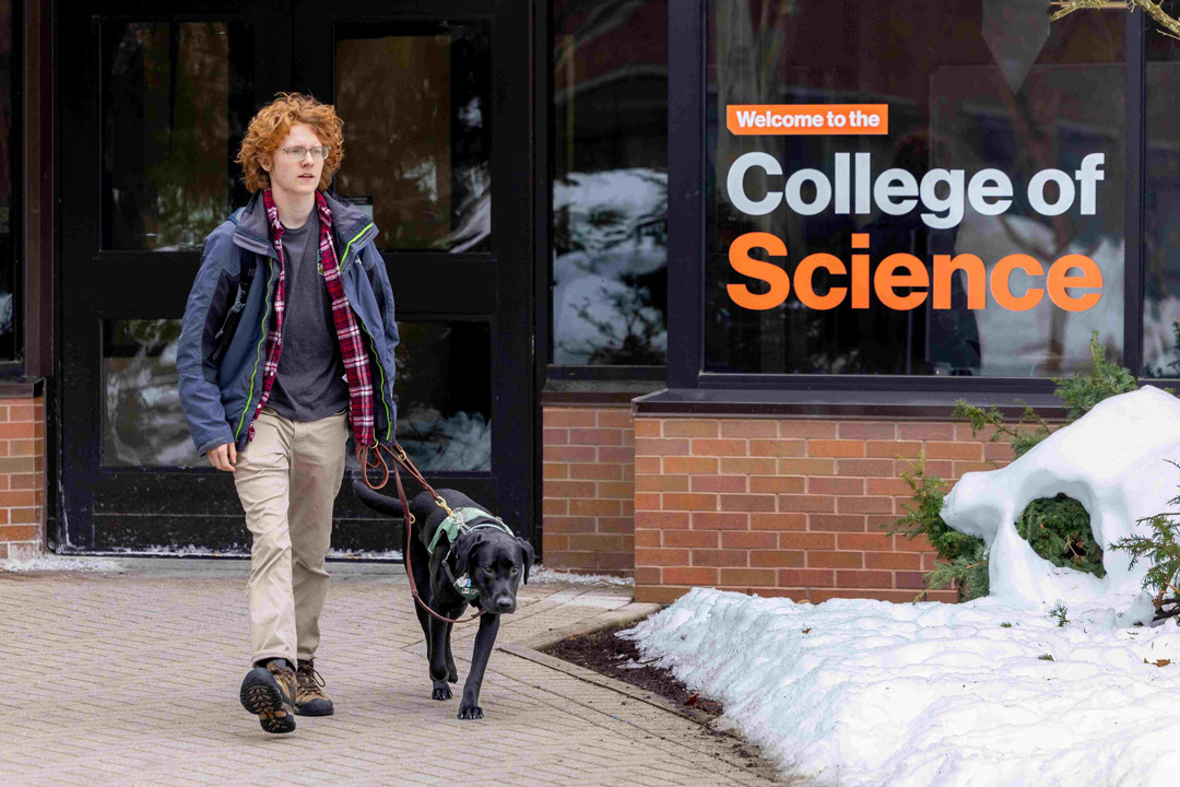 'a man with red hair walks with a black dog outside a building labeled College of Science'