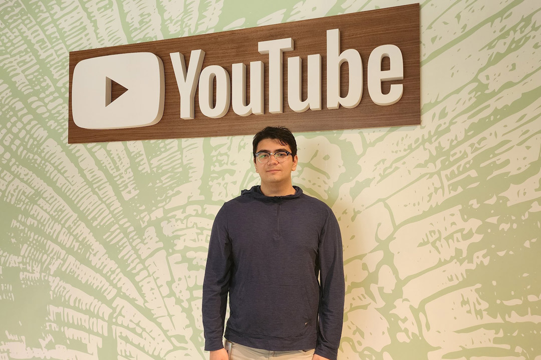 'a man with dark hair and glasses stands under a YouTube sign on a green and white patterned wall.'