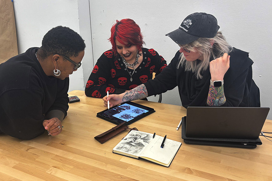 'Three students collaborate around a tablet at a classroom table, reviewing digital artwork alongside sketchbooks and a laptop.'