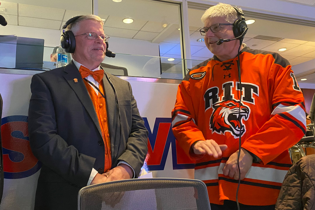 'two men sit in a press box speaking to each other with headphones.'