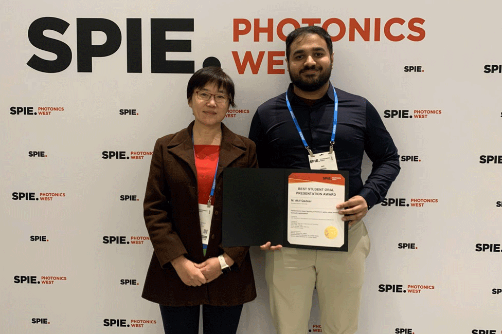 'Two people stand in front of a banner, with man on right holding a certificate.'
