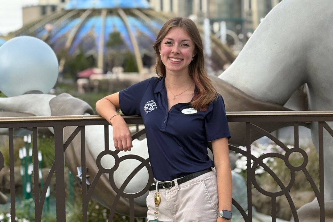 'a college age woman in khaki pants and a blue polo shirt leans against a metal railing at Universal Studios Orlando'