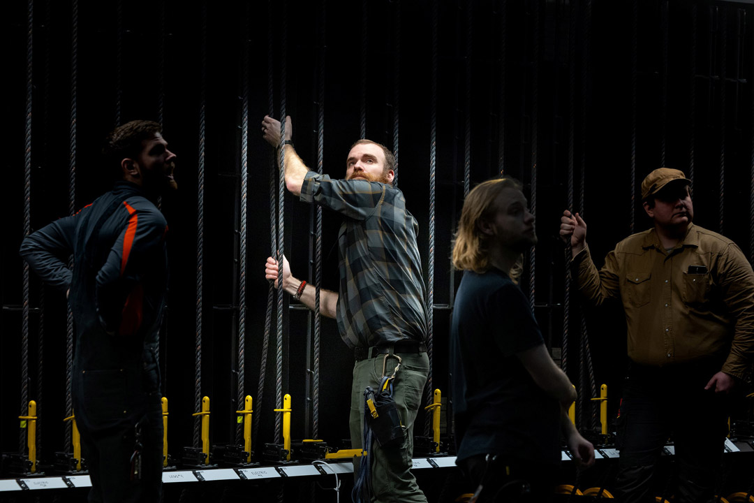 'A man shows how to lower and raise set pieces behind the scenes of a theater as students look on.'