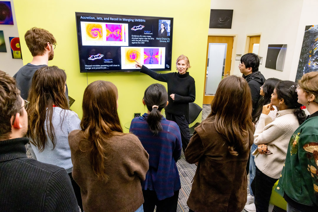 a woman with blonde hair points to a screen. in front of her, students gather to watch her speak.
