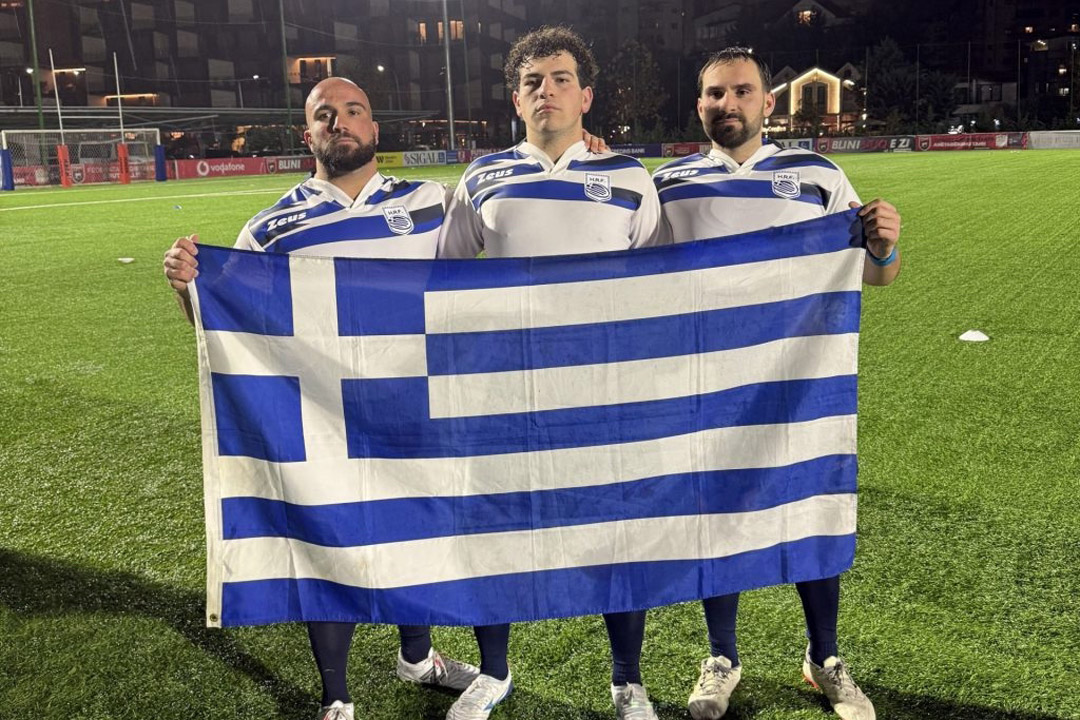 three men hold a large greek flag while standing on a soccer pitch.